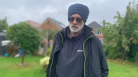 A man wearing a turban and glasses looking sad. He is dressed in a black fleece and rain coat. 