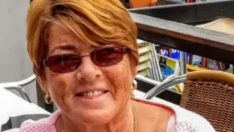 Susan Sergison smiling right at the camera. She has ginger hair swept to the left, covering her forehead. She is wearing sunglasses and a pink cardigan and has small hooped earrings in her ears. She is sitting on a wicker chair with a metal frame.