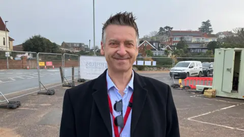 Paul Hayward, wearing a black overcoat, with a blue shirt, with sunglasses tucked into the open shirt collar. He's stood in a construction site.