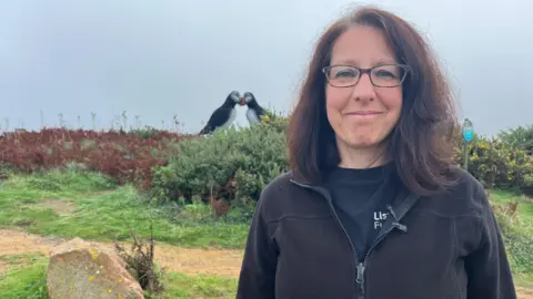 BBC Cristina Sellares - a woman with dark brown hair wearing rectangular glasses, a black top with a black thin fleece on top. In the background is a large sculpture of two puffins with their beaks touching, outside on grassland and grey skies behind.