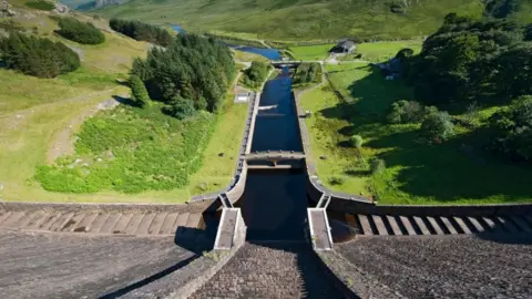 Water overflow from the dam of the Claerwen Reservoir with green hills and trees to the sides
