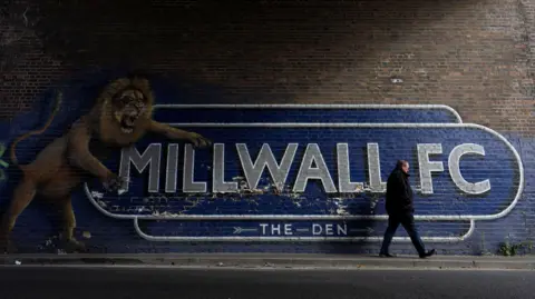 A man walks past a mural on a brick wall near Millwall's ground. Millwall FC's name has been painted in large letters on a blue background. Below that are the words "The Den" and an arrow pointing towards the stadium. The mural also features a lion, which is up on its back legs and is roaring.