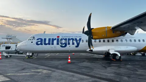 A close up side view of an Aurigny aircraft. The plane is white and yellow. Aurigny is written down the side. 