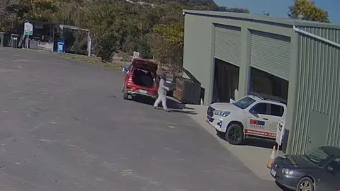 A person dressed in a grey tracksuit holding a box walks into a green shed away from red car with its boot open.