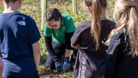 In the foreground are the backs of three children, a boy and two girls, in casual sportswear. They are standing watching a woman knelt down in the background, on grassy ground, tending to a sapling just planted into the ground. The woman is wearing a long sleeve black t-shirt, with a short sleeve green Forestry England branded polo top over the top of the t-shirt. She is wearing black trousers, blue gardening gloves, and she has brown hair tied up and is wearing glasses. It's sunny.