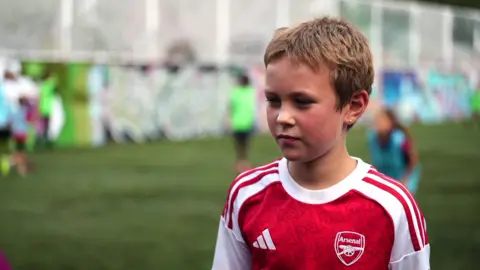 A boy in a red Arsenal football T-shirt stands on the pitch.