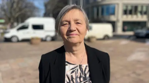 A woman wearing a black blazer over a floral top smiles at a camera. She is standing on a pavement outdoors. Parked vans are in the background behind her.
