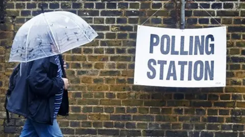 A person in a dark navy raincoat, holding a clear umbrella with musical notes on it. They are walking past a brick wall that has a white sign hanging on it. The sign reads in black "polling station"