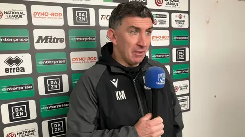 A man, wearing a grey and black jacket, holding a blue microphone and standing in front of an advertising board