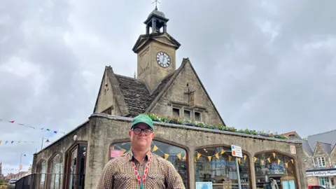 A man, wearing glasses and a green baseball cap, stand in front of a stone clocktower. Its lower section is a glazed arcade. Flowers can be seen on the lower roof and bunting laces the windows.