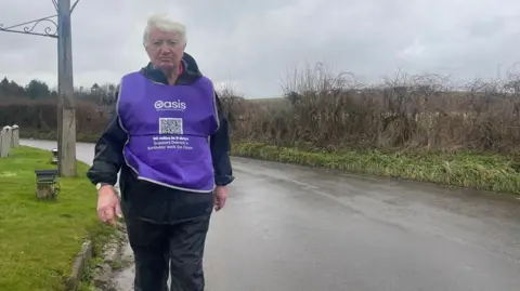 Derrick Downs wearing a mauve bib which says Oasis. He is walking along the side of a road towards the camera in wet conditions.