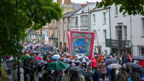 A miners' banner is carried down a street in Durham during the Miners' Gala in 2024. People with umbrellas can be seen gathered on the pavement.