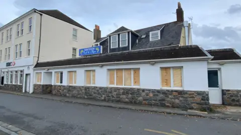 BBC Boarded up windows at the front of Northside Fish and Chips shop. The building is white and has a small blue sign located on top at the centre. There is a brick wall at the front and a door to the right.