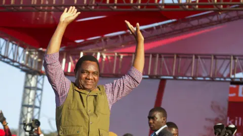 Zambia's President Hakainde Hichilema in green half-coat and black trousers lifts his hands to greet United Party for National Development supporters