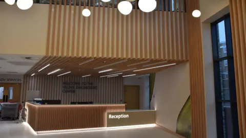 The wooded interior of the centre's reception. A bright reception desk is in the middle of the shot, with wood panelling stretching to the ceiling and the floor above.