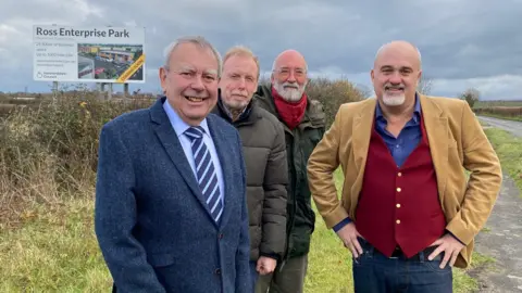 Four men standing outside in a field, with a large sign behind them that says 'Ross Enterprise Park'.