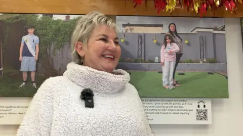 A smiling Michelle Sank. She's wearing a white fleece top, stood in front of two of her photographs. 