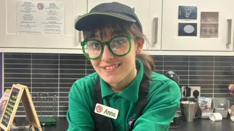 Anna, a teenage girl with green framed glasses in the kitchen. She is smiling and wears a green T-shirt.