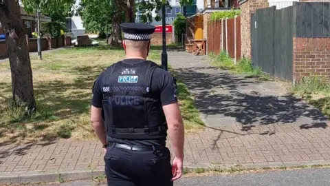 Police officer wearing black shirt and trousers and black cap with white and black braiding. The word POLICE is on the back of the shirt. He has his back to the camera and is walking on a block-paved path towards a group of two-storey terraced houses. There is a grassed area immediately in front of him and a tree, the branches of which are hanging above the pavement.