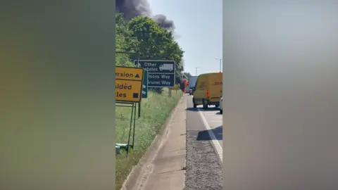 A road with a number of vehicles on it including a yellow van. In the distance is a fire with a large plume of smoke above it. On the right hand side of the road are road signs. 