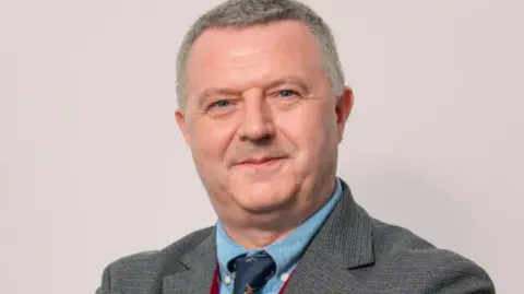 Kevin Donaghy, with grey hair, grey suit, blue shirt and blue tie.