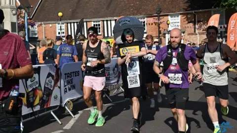 Dan Hudson Participant wearing a large helmet-shaped costume and a Marie Curie charity bib walks among runners during a road race in a town centre. Spectators line the route behind barriers displaying sponsor signage, with brick buildings and event banners visible in the background.