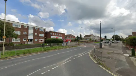 A generic Google image of Middle Park Way -  a wide junction with footpaths  with a three-storey block of flats opposite, a convenience store and houses further down the street