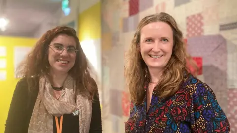 Kirsty Hartsiotis from Swindon Museums and Anna Bryant from the Wessex Museums partnership look into the camera standing in front of a quilt.