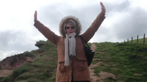 A woman in her seventies is pictured with her arms in the air. She is wearing a brown coat with a fur-lined hood pulled up over her head and a grey scarf. She is standing on a hill with a path rising up behind her 