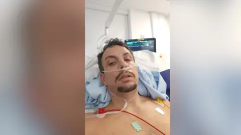 Leandro Varela Dominguez lies in a hospital bed. He is shirtless and connected to monitors and a nasal canula for oxygen. He has short brown hair and a short beard and moustache. A monitor screen is visible in the background. 