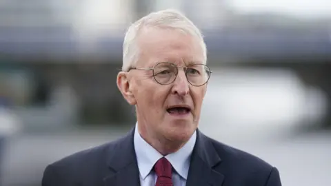 PA Media A man with white hair and black circular glasses wears a dark suit jacket, a light shirt and red tie