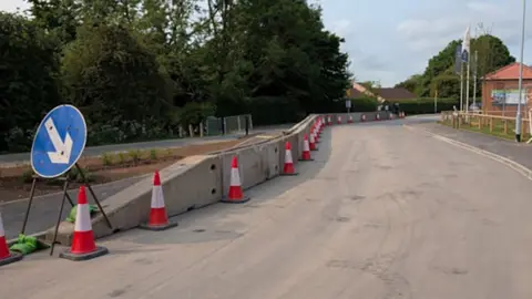 Concrete crash barriers along the side of a road with orange and white traffic cones in front of it and a blue road sign with arrow