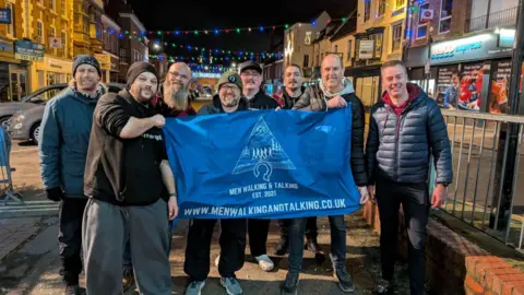 Men in a walking group stand in a shopping street and hold up a banner. It is night-time and there are Christmas lights above the road. Several men are standing near railings and they are wearing jackets, hats and trainers.