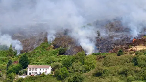 White smoke coming out of fires on a green valley. There is a white house with a terracotta roof in the foreground of the photo
