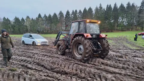 Mel Slade A tractor pulling a silver car over badly churned muddy ground, with a row of trees in the backgroud.