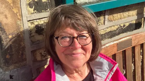Grace Wood/BBC A woman sitting on a bench with round glasses and a short brown bob. She is wearing a pink rain jacket.