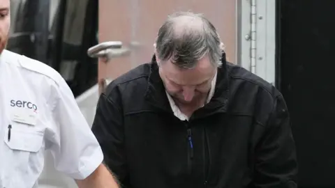 Ian Silvester being led from a custody vehicle by an officer, with his head bowed. The officer is partially in the picture on the left side of the image. 