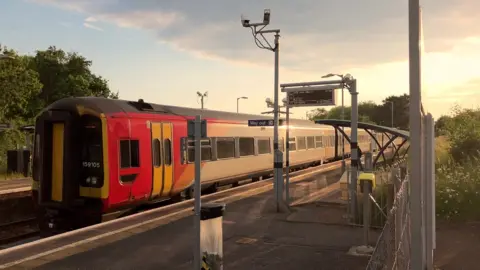 A South Western Railway train travelling through a station past an empty platform on a sunny day.
