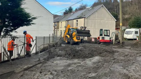 BBC A street in Cwmtillery covered in black slugged piled to make it impassable. Two men in high vis orange jumpsuits and hard hard carry shovels as they stand on the pavement. A yellow digger with a large scoop and a smaller white and red digger with a small scoop are working to move the debris 