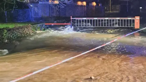 London Fire Brigade Road with water flowing down it. A barrier has water gushing underneath it. It is night time