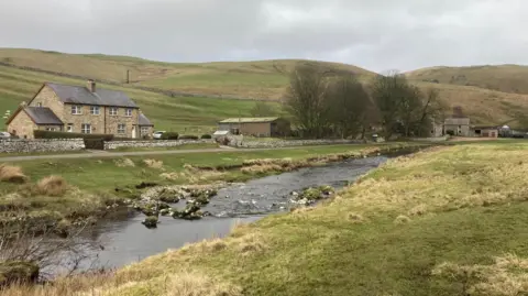 Sam Wood Two houses are on the left with a small river in front. To the right and in the distance behind trees are more farm buildings. They all sit at the foot of a number of hills. 
