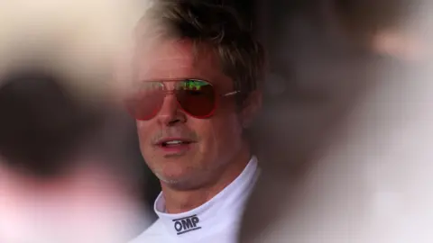 Image shows Hollywood actor Brad Pitt. He has short blond hair, is wearing shades, and a white polo neck top. 