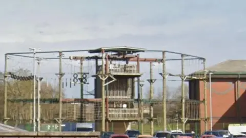 A number of wooden poles form the structure of the high ropes course, with ropes and netting seen and a multi-level tower structure at the front. Some trees can be seen in the background, with part of the sports centre building also visible on the right. 