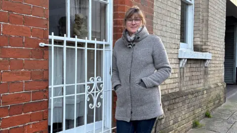 Supplied Tammie McNeill, wearing a grey coat, scarf, glasses and denim jeans, standing next to the only window of her flat that has not been damaged. It’s a long glass window with a white frame and a white, metal window guard made up of bars on top of it.