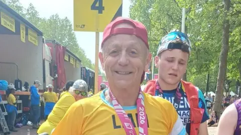 Liam McHugh Liam is wearing a read peaked cap, which he has turned back--to-front. His jersey is yellow and he has a pink ribbon around his neck, from which presumably hangs a medal. 