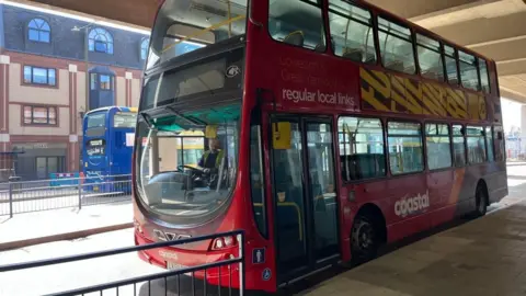 A red Coastal Reds bus, stopped at Great Yarmouth bus station. The bus is a double decker, painted in red, orange and burgundy, with "regular local links" as a promotional slogan and an advert on the side. On the far side of the station is a blue bus, facing in the opposite, southbound, direction.