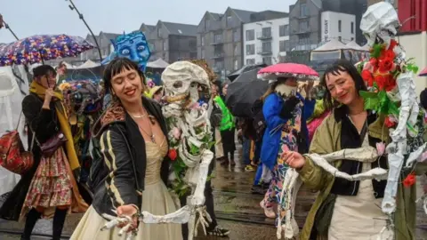 Bristol Experimental Puppet Festival A crowd of people are standing outside in the pouring rain, dancing with homemade skeleton puppets. People in the background are holding umbrellas.