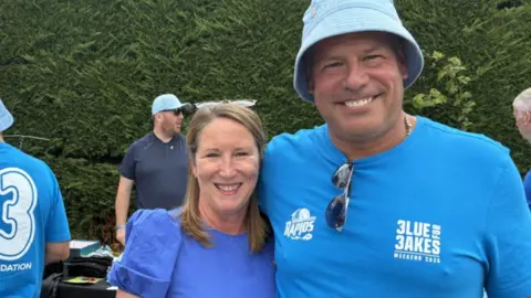 A man and woman wearing blue t-shirts smile at the camera with their arms around each other