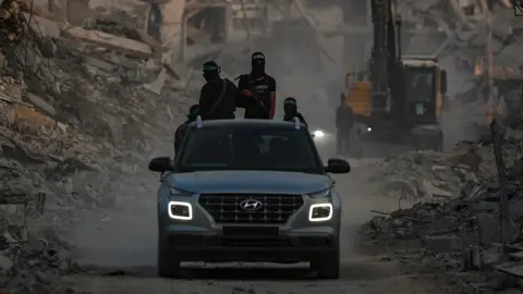MOHAMMED SABER/EPA/Shutterstock Fighters of the Al-Qassam Brigades, the military wing of the Hamas movement, wearing balaclavas and headbands, and carrying guns, stand guard behind a car as they search for the bodies of Israeli hostages alongside Red Cross workers in Al Shejaeiya neighbourhood in the east of the Gaza City, Gaza Strip. A bulldozer is seen among rubble in the background.