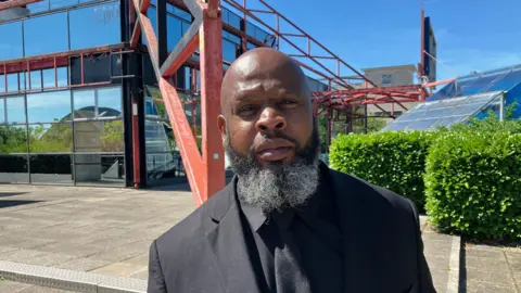 Sam Read/BBC Jason stands outside The Point wearing a black shirt, tie and suit jacket. He has a grey-black beard and is squinting slightly at the camera in the sun. The sky is blue behind him and the Point's mirrored glass reflects surrounding trees and buildings.